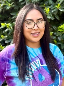 Smiling woman in tie-dye shirt and glasses standing in front of greenery